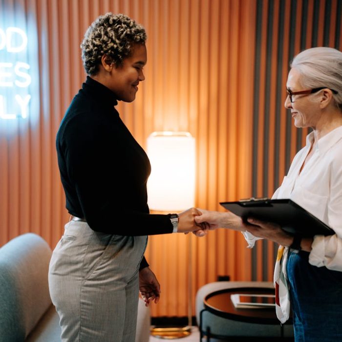 Two diverse businesswomen in a stylish office shaking hands under a 'Good Vibes Only' sign.